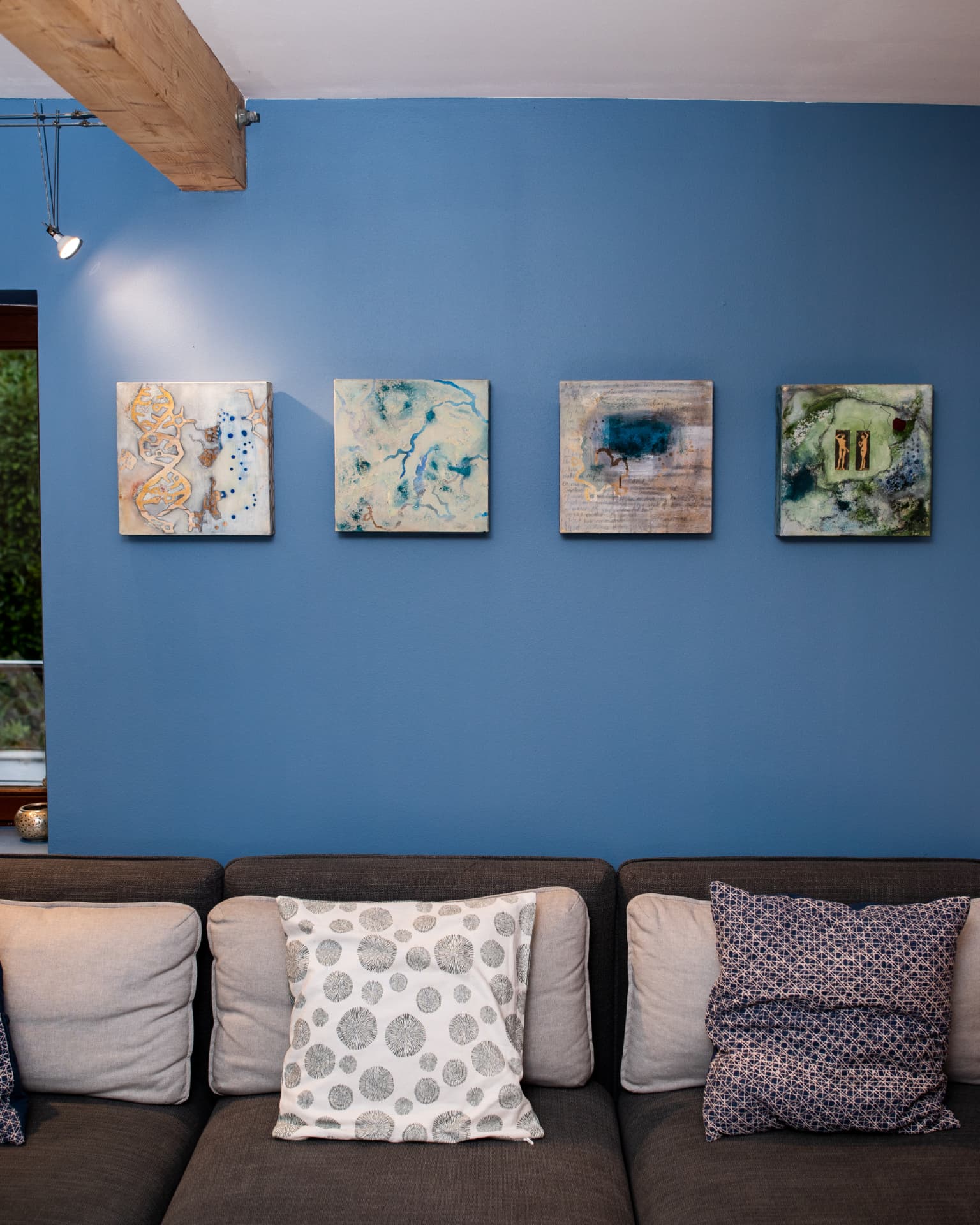 The four Origins of Life paintings hanging in an even row on a blue wall above a grey sofa, under a wooden ceiling beam