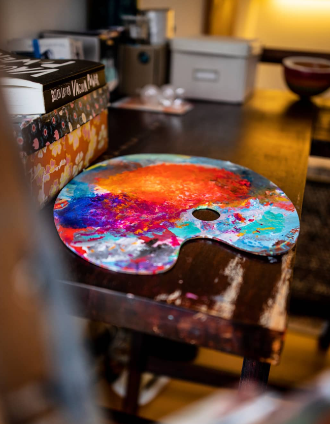 A thumbhole palette resting on a dark wooden table covered in blends of orange, magenta, turquoise and blue paint